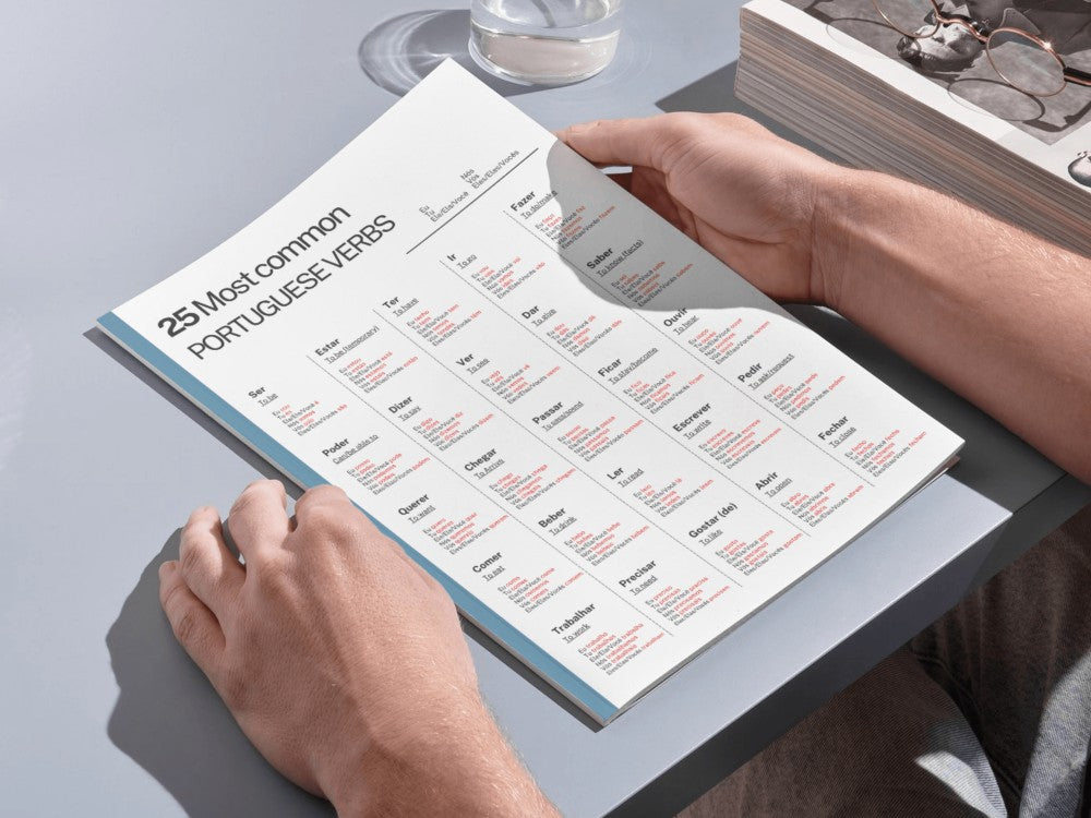 Person holding a tablet displaying a Portuguese verb chart on a light gray surface.