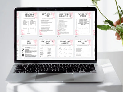 A laptop on a white surface displaying a digital game or activity sheet set with pink floral accents and black text. A hand holding green leaves is visible on the right side.