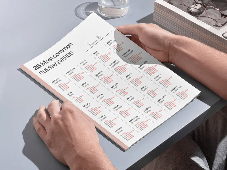 Person holding a book with Russian verb conjugation charts on a light gray surface.