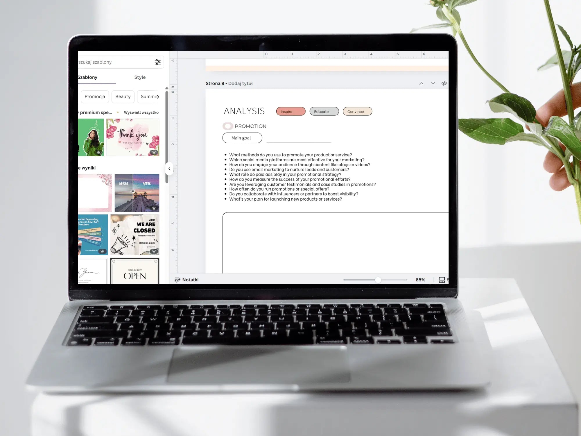 A silver laptop on a white surface displaying a digital design or marketing interface, with a hand holding green leaves on the right side of the frame.