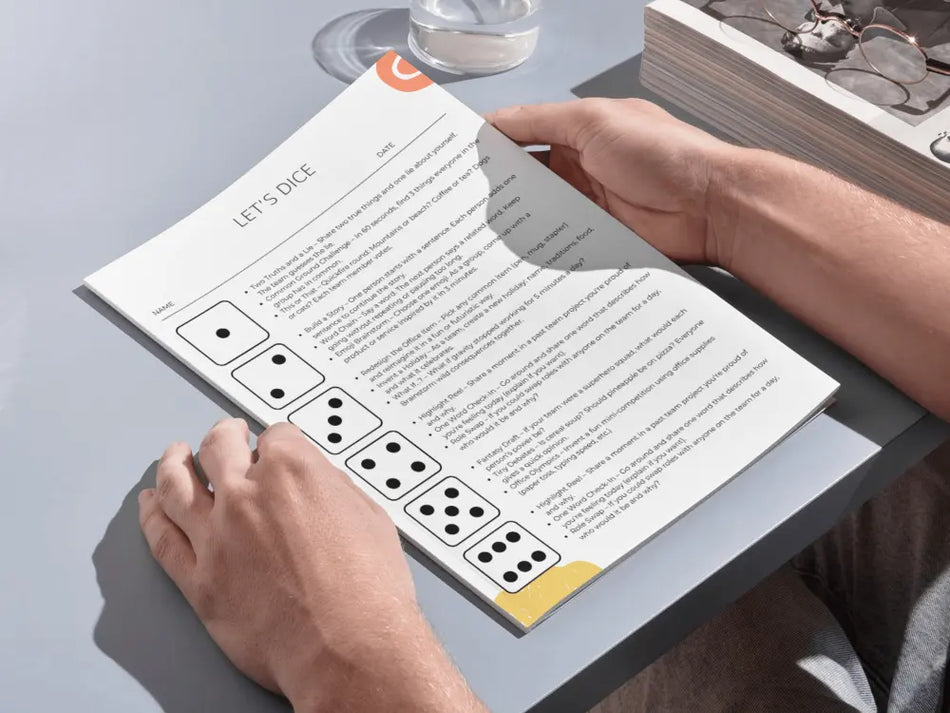 A person holding an open printed booklet or card on a gray tabletop. The page shows the heading 'LET'S DICE' with text and several black-and-white dice illustrations along the left side.