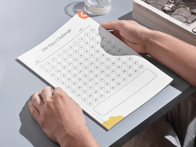 Close-up of a person’s hands holding a printed version of the 100-day challenge sheet on a clean grey desk, demonstrating the physical, tactile use of the progress log.