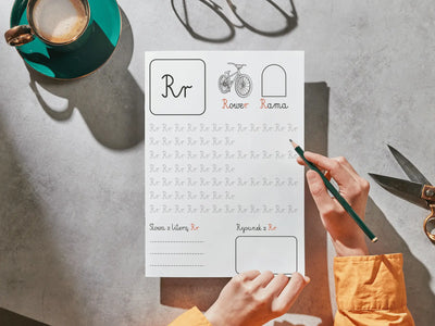 A person's hands hold a printed Polish handwriting practice sheet for the letter 'Rr', using a green pencil. The sheet features a boxed 'Rr' at the top, simple line drawings, and rows of cursive tracing exercises.