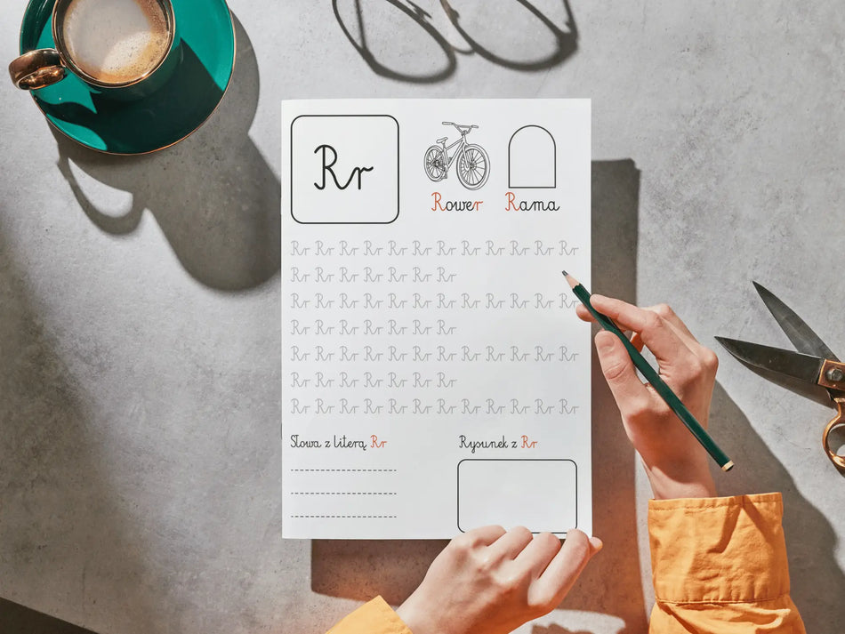 A person's hands hold a printed Polish handwriting practice sheet for the letter 'Rr', using a green pencil. The sheet features a boxed 'Rr' at the top, simple line drawings, and rows of cursive tracing exercises.