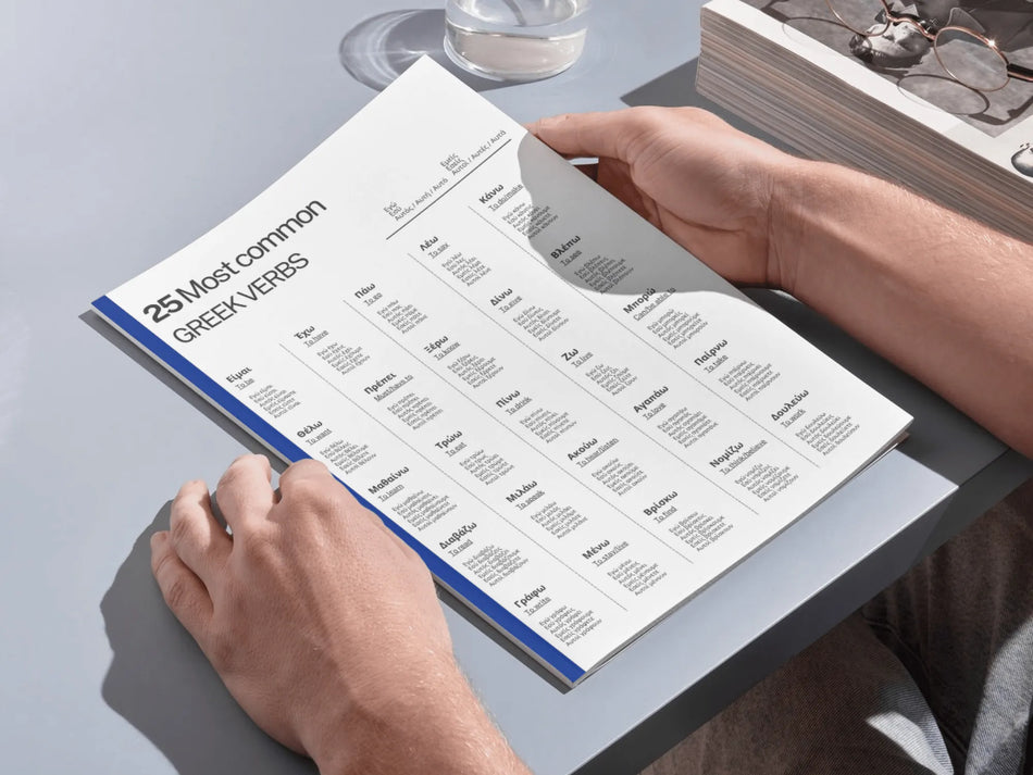 Person holding a book titled '25 Most Common Greek Verbs' on a table.