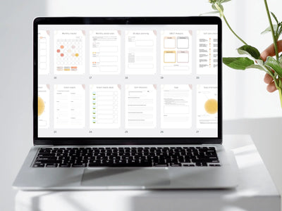 A close-up view of a laptop screen on a white desk showing a grid of 10 different planner pages including vision boards, self-reflection prompts, and goal prioritization charts